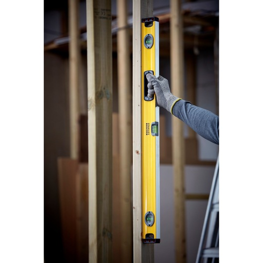 STANLEY 1-43-536 spirit level held vertically against wooden beam, gloved hand showing usage on construction site.