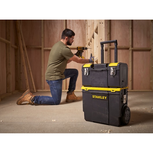 Photo of STANLEY 1-70-326 rolling tool box with yellow accents, shown on jobsite with worker in background.