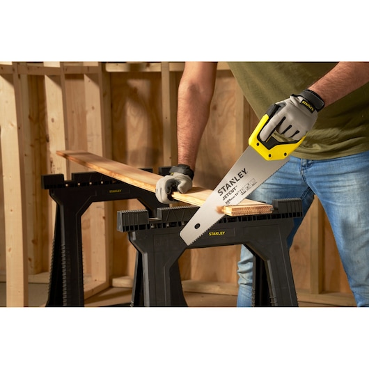 STANLEY 2-15-281 hand saw with yellow handle cutting wood plank on black saw horse in workshop.