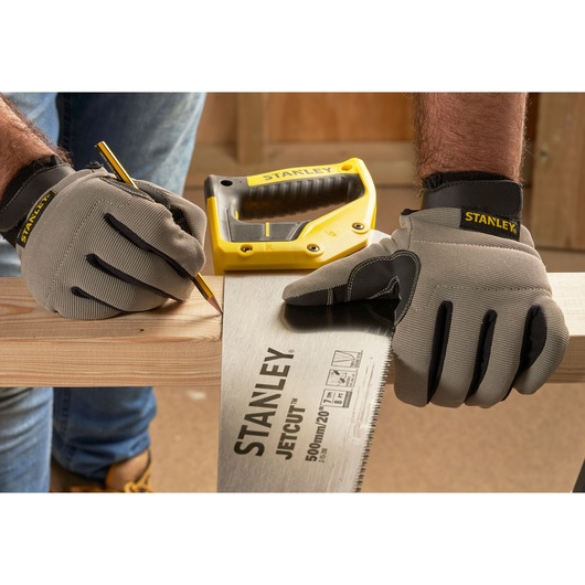 STANLEY 2-15-288 hand saw and yellow stapler on wood, person with gloves marking cut line, workshop background.