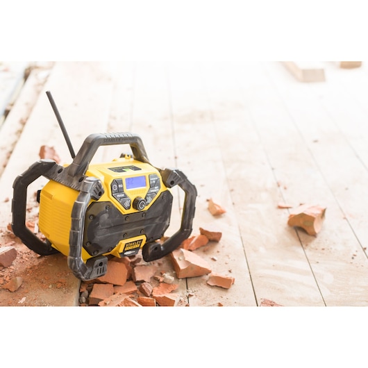 STANLEY FMCR001B jobsite radio with black protective frame on wood floor surrounded by broken bricks, front angle.