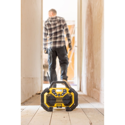 STANLEY FMCR001B jobsite radio with yellow and black design on a wooden floor, worker blurred in background.