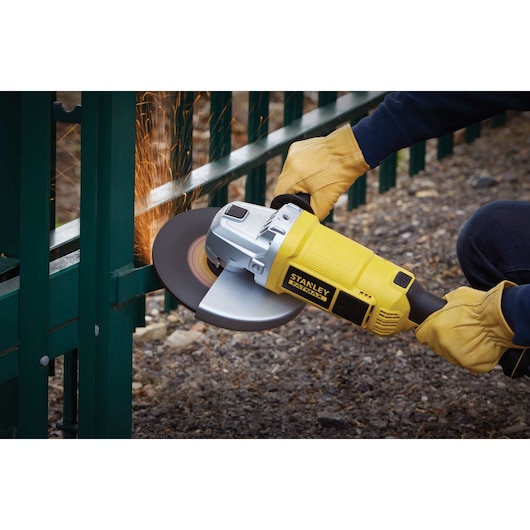 STANLEY FME841 angle grinder in use, yellow body, grinding metal fence, side view, sparks flying.