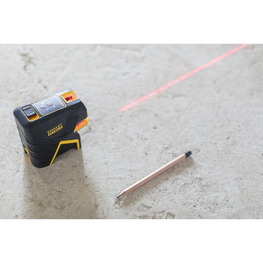 STANLEY FMHT1-77415 cross line laser level with visible red laser line and pencil on concrete floor, angled view.
