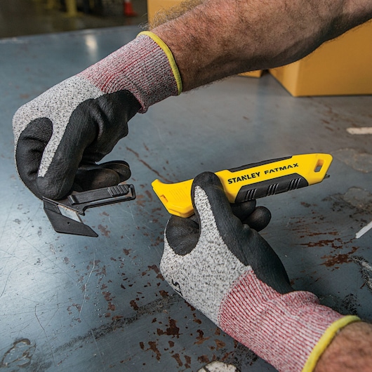 Photo of STANLEY FMHT10358 FATMAX utility knife with yellow handle held by gloved hands on industrial surface.