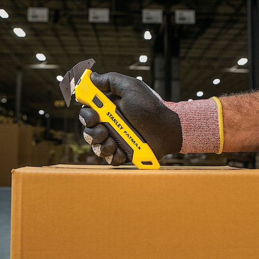 STANLEY FMHT10358 utility knife with yellow handle, held in gloved hand, cutting cardboard box.