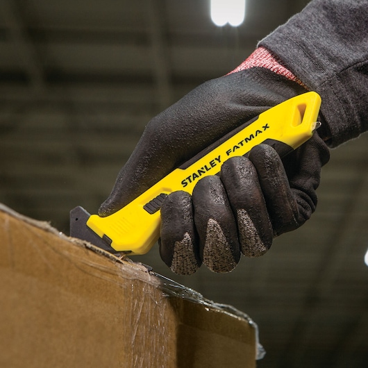 Stanley FMHT10358 utility knife cutting cardboard, close-up, yellow handle, gloved hand, photography.