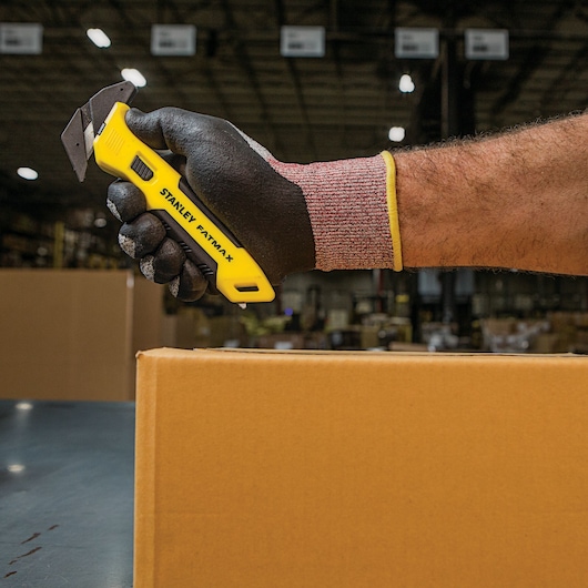 Photo of STANLEY FMHT10358 utility knife in gloved hand, cutting a cardboard box in a warehouse.