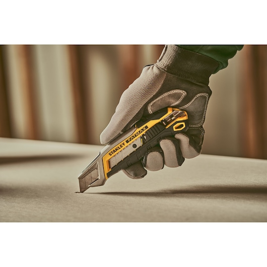 Stanley FMHT10594 utility knife held by gloved hand cutting material, photography close-up.