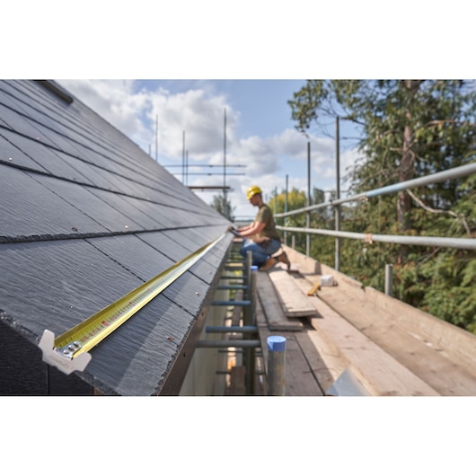 STANLEY FMHT38232-0 tape measure extended on roof, construction site with worker and scaffolding, outdoor use.