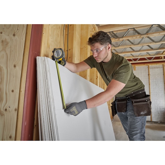Photo of a worker measuring drywall with a STANLEY FMHT38232-0 tape measure in a wooden framed room.