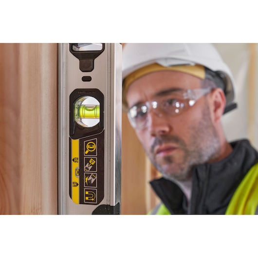 STANLEY FMHT43671-1 spirit level with yellow accents, vertical view next to wood, blurred worker in background.
