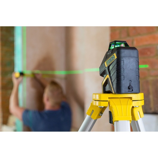 STANLEY FMHT77617-1 green laser level on tripod projecting line across brick wall, worker measuring in background.