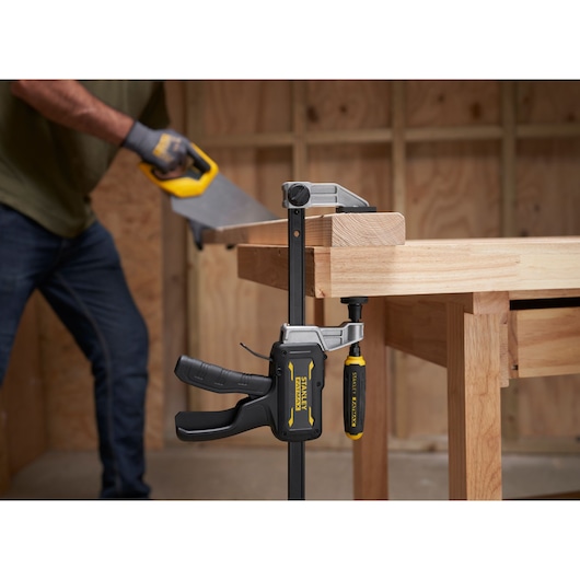 STANLEY FMHT83006-0 clamp securing wood on workbench, person sawing wood in workshop.