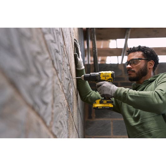Person using STANLEY SFMCD715D1K cordless drill on a brick wall, wearing gloves, indoor construction setting.