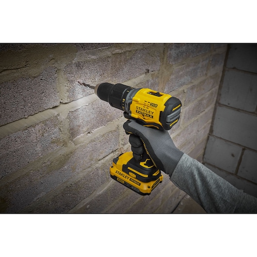 Stanley SFMCD715D2F cordless yellow drill used on brick wall, close-up of tool in gloved hand.