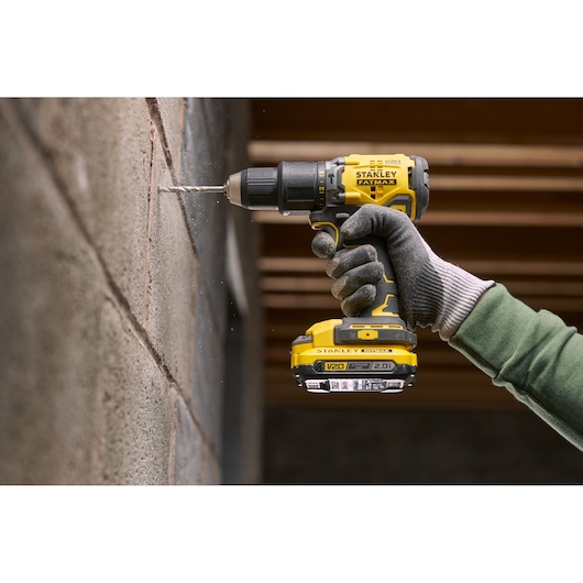 STANLEY SFMCD715D2K cordless drill, yellow and black, drilling into masonry wall, side angle.