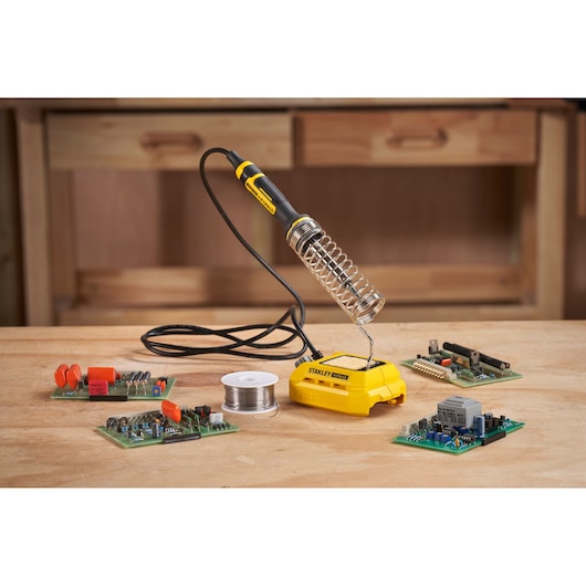STANLEY SFMCE040B soldering iron on yellow base with circuit boards and solder wire on workbench, front angle.