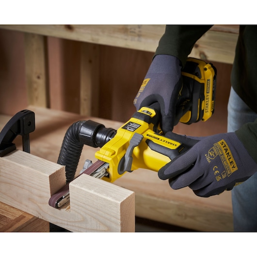 Photo of STANLEY SFMCE210D2 cordless belt sander in use on wood, close-up of hands wearing gloves.