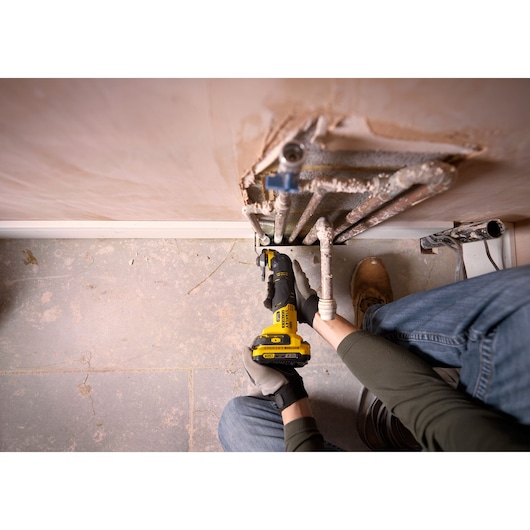 Photo of STANLEY SFMCE500B oscillating multi-tool in use cutting pipes near a wall, user kneeling on floor.