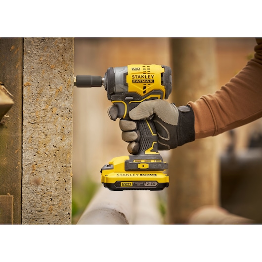 Photo of STANLEY SFMCF910B impact driver in use, yellow and black design, cordless, gloved hand operating tool outdoors.