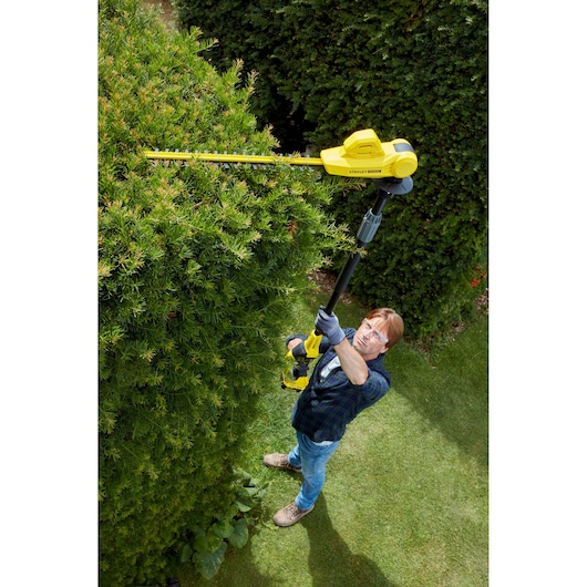 STANLEY SFMCPH845B photography of cordless pole hedge trimmer in use trimming tall green bushes.