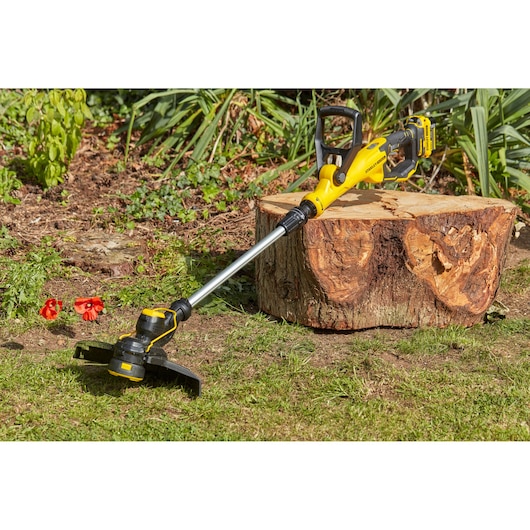 STANLEY SFMCST933B cordless grass trimmer in yellow and black lying on grass near a tree stump outdoors.