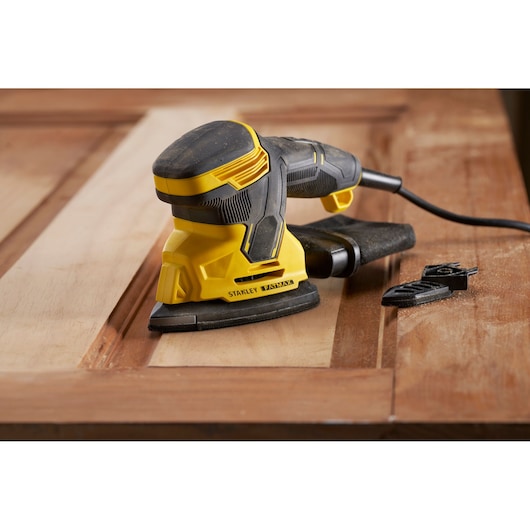 STANLEY SFMEW210S sander in use on wooden surface, close-up side angle, with attachments nearby.