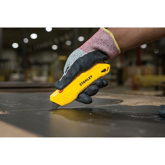 Photo of gloved hand using STANLEY STHT10368-0 yellow utility knife to cut black material, close-up.