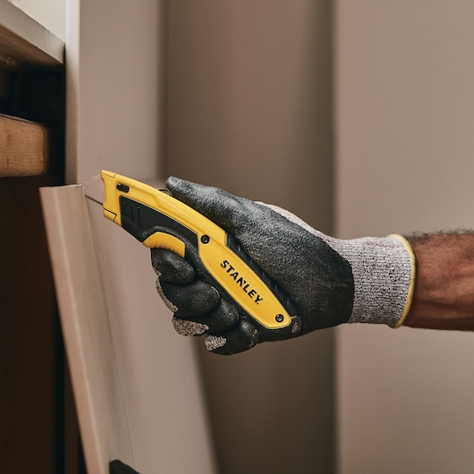 Stanley STHT10479-0 utility knife held in gloved hand, cutting a panel. Photography product category.
