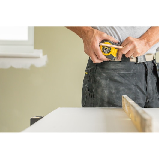 STANLEY STHT36803-0 tape measure held by worker in dusty work trousers beside white table.