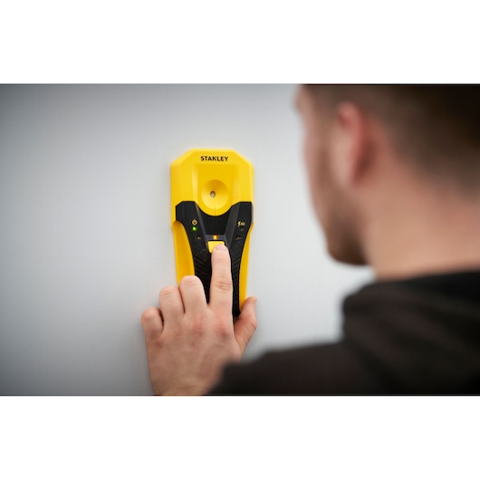 STANLEY STHT77588-0 stud finder device in use against a wall, photography showing hand pressing button.