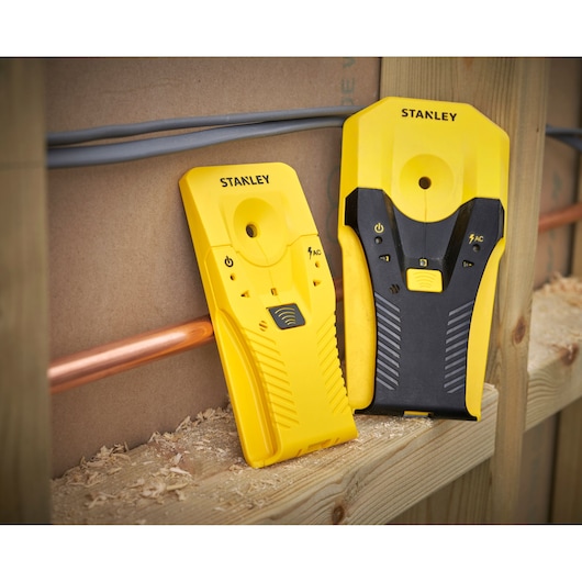 Two yellow STANLEY stud finders on a wooden surface near pipes and wires during construction.