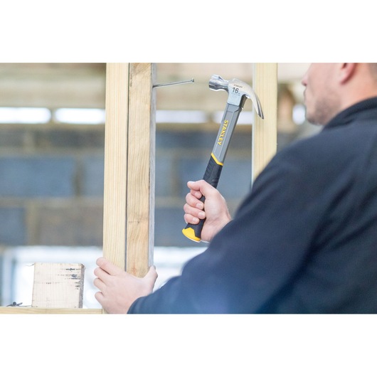 Stanley STHT9-51309 hammer in use, person driving nail into wood frame, close-up scene.