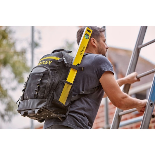 STANLEY STST83307-1 black tool backpack with yellow STANLEY level in side pocket, shown outdoors on person climbing ladder.