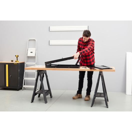 Photo of STANLEY STST97599-1 workbench in workshop, man assembling black metal frame on wood surface.